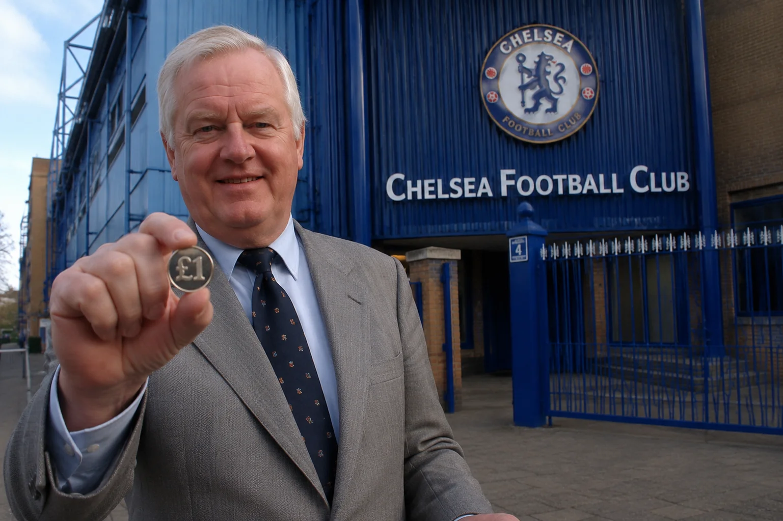 Former Chelsea chairman Ken Bates smiling while holding up a one-pound coin (£1) in the foreground, standing directly outside the entrance gates of Stamford Bridge stadium underneath a large Chelsea Football Club sign and crest.