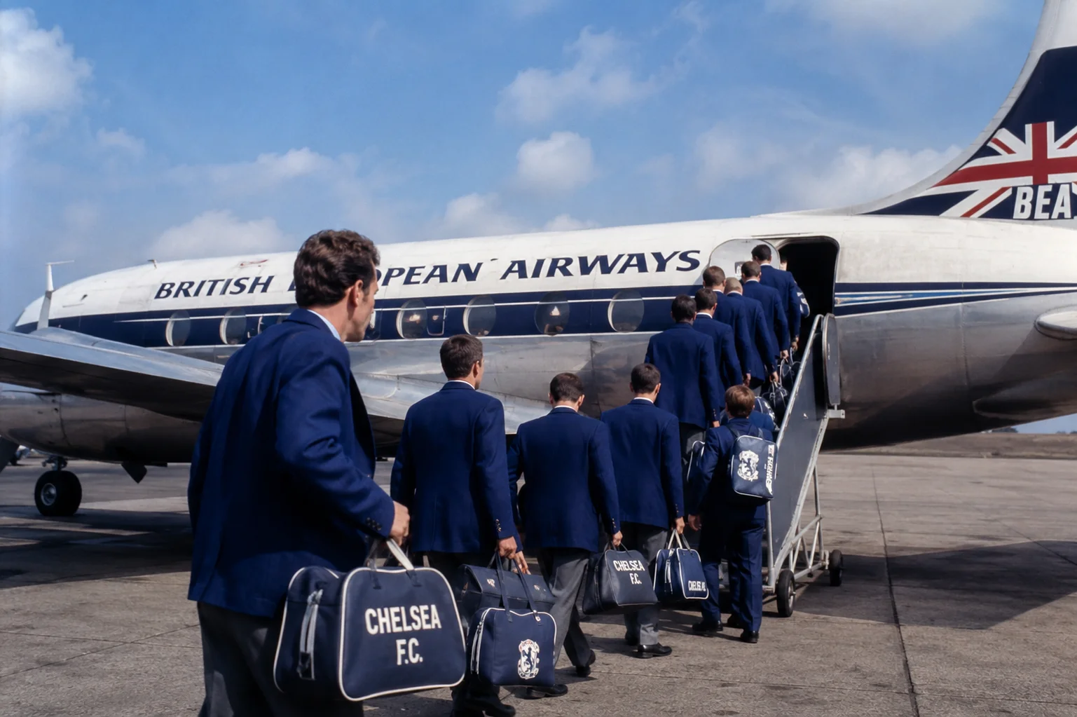 Chelsea football players wearing blue club blazers and holding matching travel bags as they walk up the boarding stairs onto a British European Airways airplane on the tarmac for a match abroad.