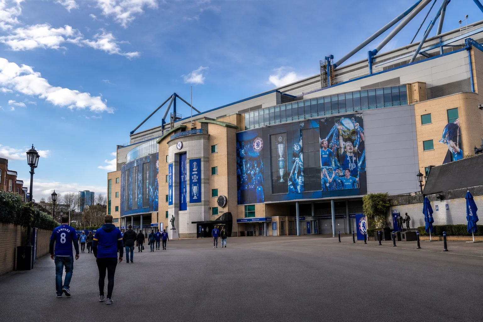 Supporters walking along the paved entrance path outside Stamford Bridge stadium, which is decorated with large blue banners showing iconic Chelsea trophy celebrations.