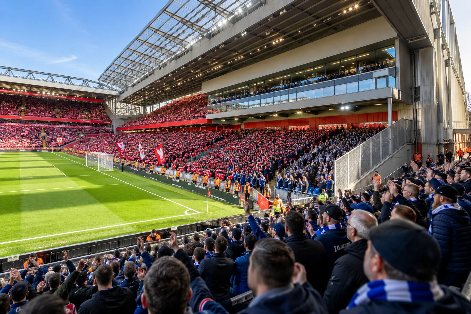 View from inside the visitor section at Anfield, showing fans wearing blue scarves and jackets clapping and cheering from their corner section, looking towards the main home stands across the green pitch.