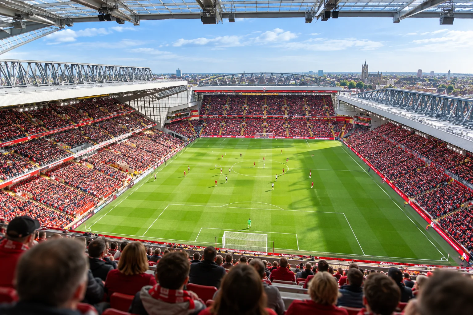 High-angle, panoramic photograph of Anfield Stadium taken from the top upper tier, showing the entire green pitch below, surrounding full grandstands, and the Liverpool city skyline in the distance under a blue sky.