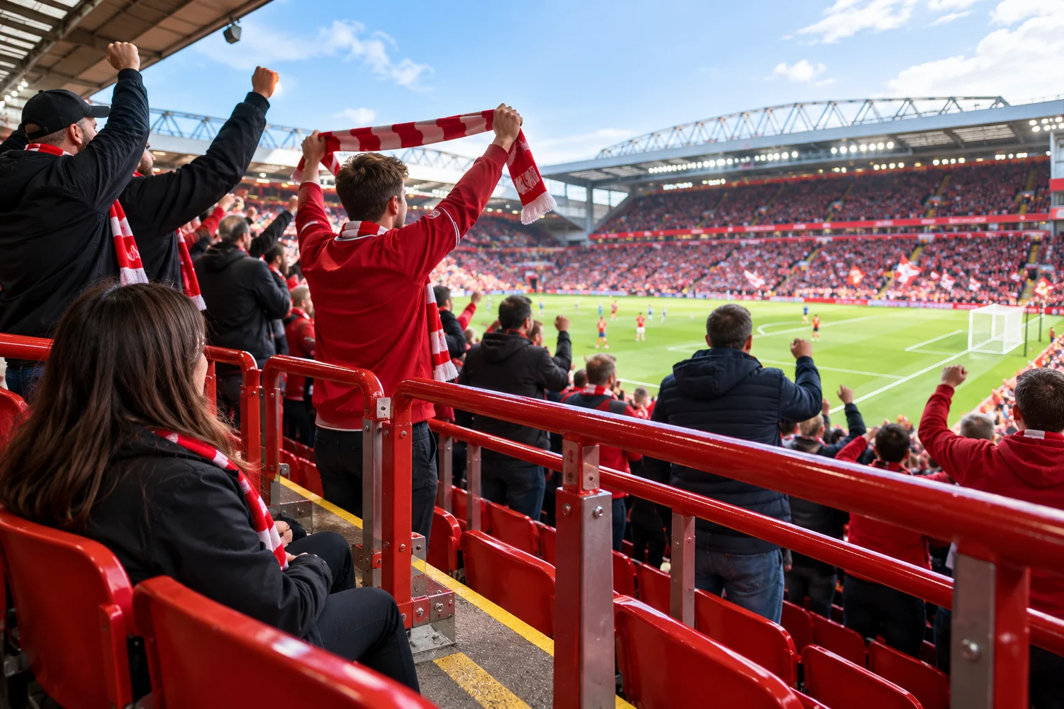 First-person perspective from behind red safe-standing rails in the stands at Anfield Stadium. Fans in red Liverpool jackets are cheering and holding up a scarf overhead, looking down towards players on the pitch.