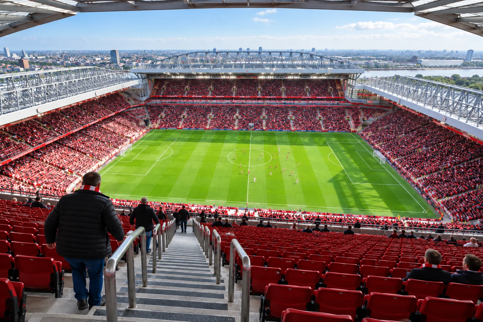 High-tier view looking straight down the central concrete aisle stairs and metal handrails towards the pitch, showing thousands of red seats filled with spectators and the River Mersey visible in the far background skyline.