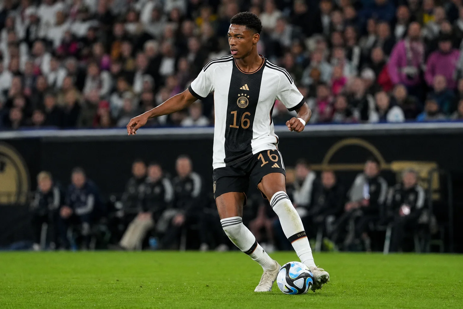 صورة للاعب الوسط الألماني عسان أوييدراوغو خلال جلسة تدريب أو مباراة. يظهر من الصدر إلى الأعلى، وينظر للأمام بتعبير جدّي وتحليلي. يرتدي الزي الأبيض الوطني لألمانيا والذي يتضمن نمط مقعد متدرج من الأسود إلى الأحمر، بينما يبقى خلفه خلفية ملعب داكنة جليدي.