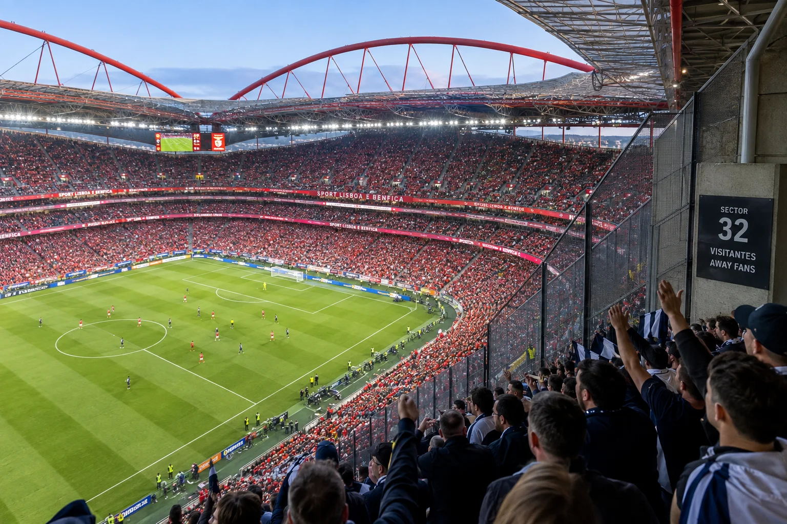 View from the away supporters' section inside S.L. Benfica's Estádio da Luz during a match. Away fans are standing behind a protective glass and mesh barrier next to a sign labeled "Sector 32 - Visitantes / Away Fans," overlooking a full stadium of red seats and the pitch.