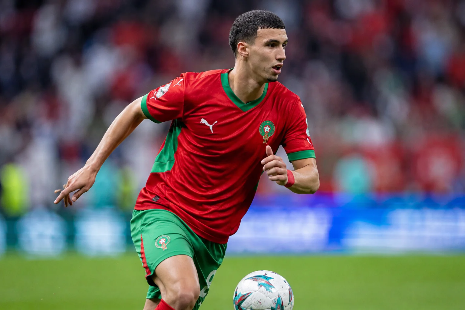 صورة عن الملعب للاعب الوسط المغربي بلال الخانوس خلال مباراة. يظهر من الصدر إلى الأعلى، يبدو مركزًا ويوجه عينيه نحو الجانب الأيسر من الملعب. يرتدي الزي الوطني الأحمر للمغرب مع تفاصيل خضراء على الكتفين وشعار الاتحاد الملكي لكرة القدم على الصدر الأيسر. تُظهر الخلفية ملعبًا مهتجًا وإعلانات تحت إضاءة ساحة ساطعة.