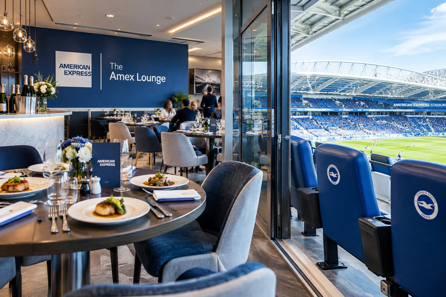 The Amex Lounge dining room at American Express Stadium, featuring round tables with plated gourmet meals, branded blue seating overlooking the stadium pitch, and a wall display with "The Amex Lounge" logo.