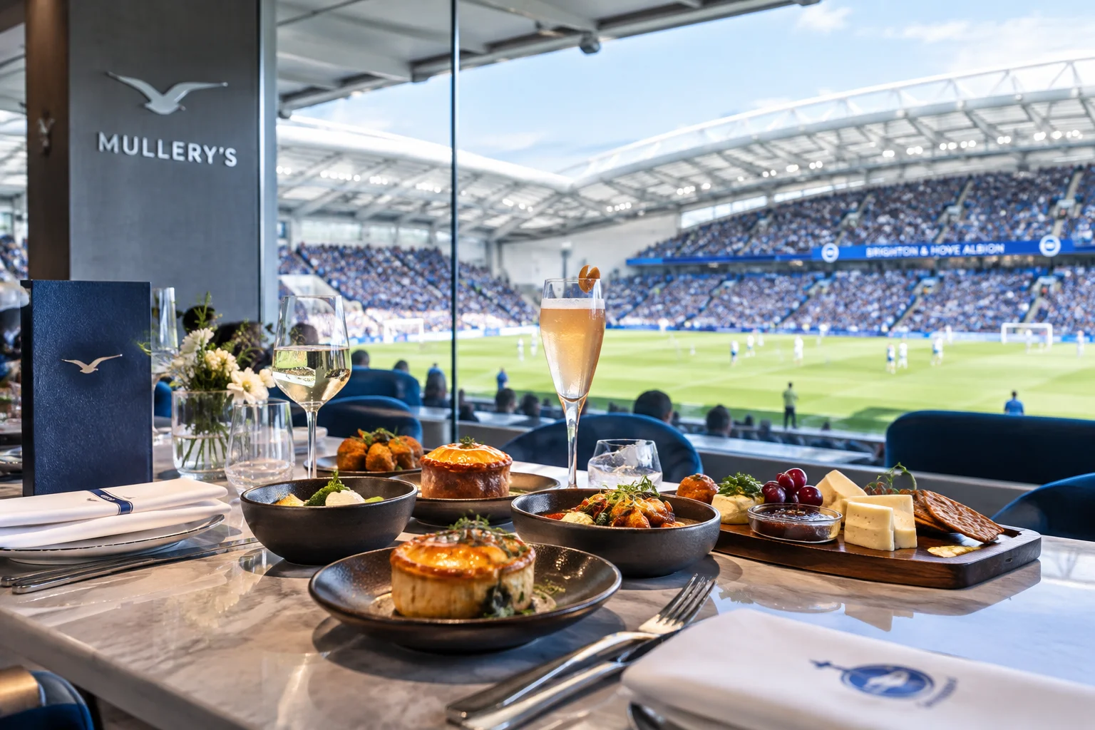 A restaurant table at Mullery's set with savory pies, appetizers, and champagne drinks directly in front of large windows overlooking a packed stadium crowd during a match.