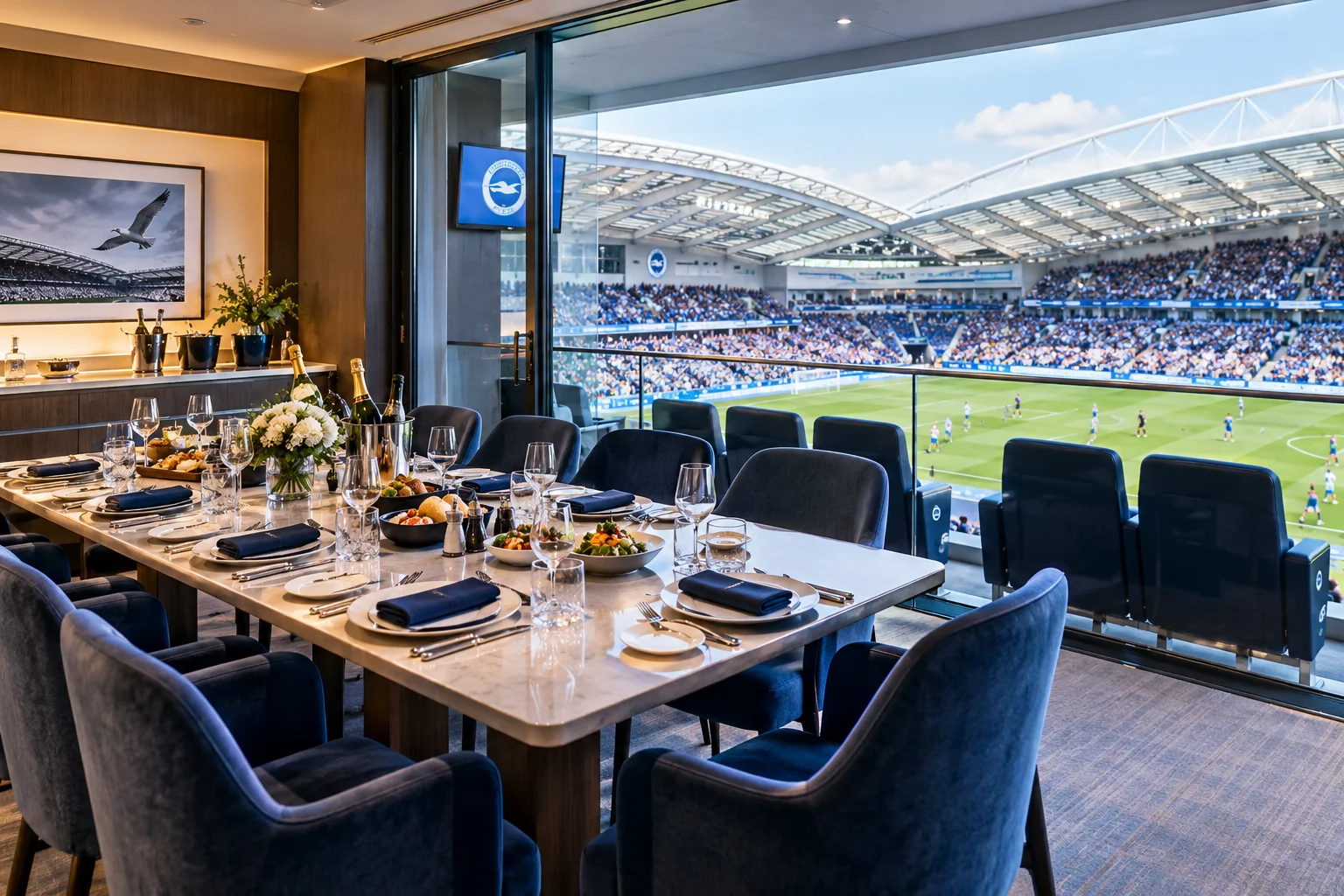 A private executive box set for dining with a long table, champagne, and appetizers, featuring floor-to-ceiling glass doors opening directly onto a view of the stadium pitch during a football match.