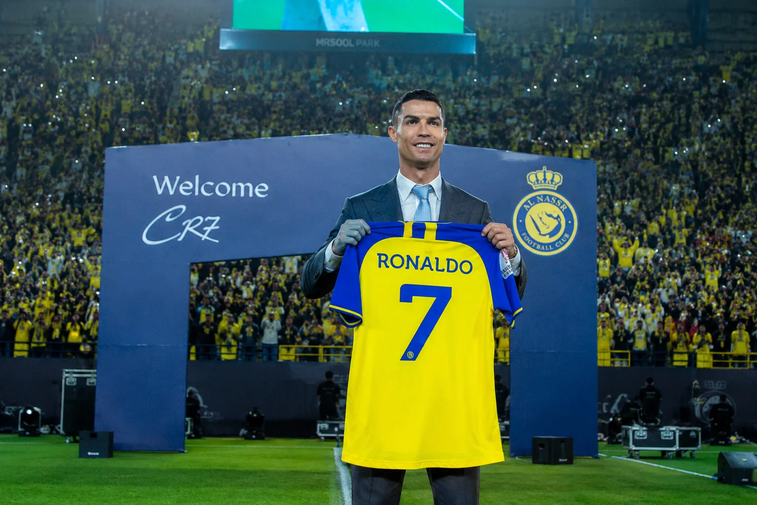 An infographic tracking Cristiano Ronaldo's historic move to Asian football with Al Nassr FC. The left panel showcases a smiling Ronaldo presenting his vibrant yellow and blue Al Nassr jersey emblazoned with his signature number 7 on the back. The background features a glowing golden graphic representation of Al-Awwal Park stadium in Riyadh under a night sky. The right panel describes his landmark signing in late 2022, his impact on the Saudi Pro League, and his prolific calendar-year scoring records in the Middle East.