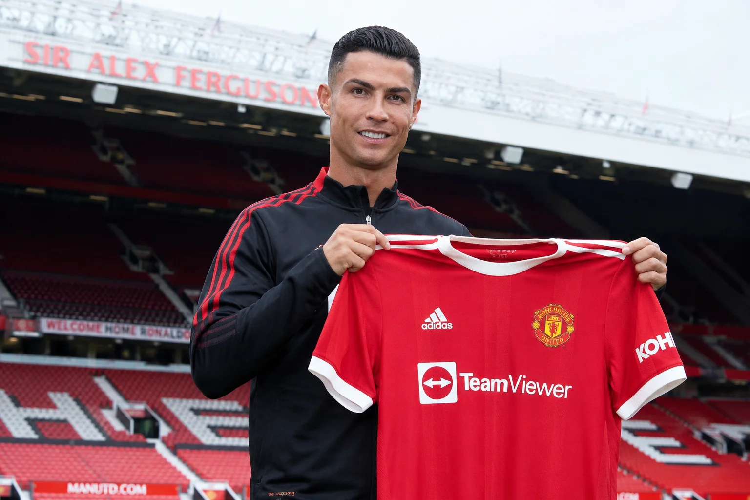 An infographic highlighting Cristiano Ronaldo's highly anticipated emotional homecoming to Manchester United in 2021. The left view shows an older, mature Ronaldo clapping toward the fans in the modern red TeamViewer-sponsored Manchester United home jersey. The background includes a dark silhouette of the Old Trafford stands illuminated by soft red accent lighting. The right view tracks his 2021-2022 season performance, detailing his crucial late Champions League group stage goals, hat-tricks against Tottenham and Norwich, and finishing as the club's top goalscorer.