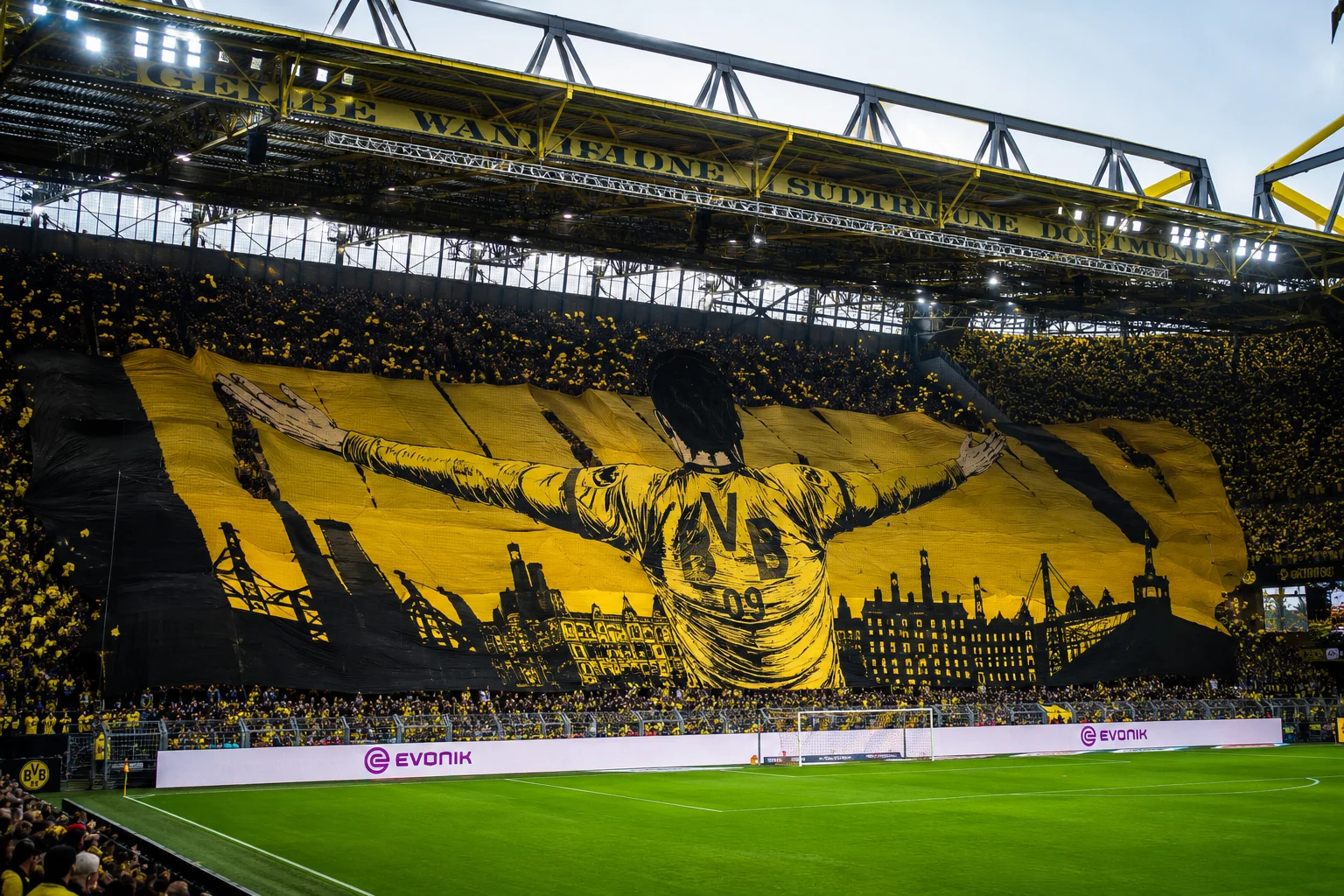 A massive, iconic fan tifo banner spanning the Südtribüne, featuring a player in a BVB jersey with outstretched arms set against the skyline of Dortmund.