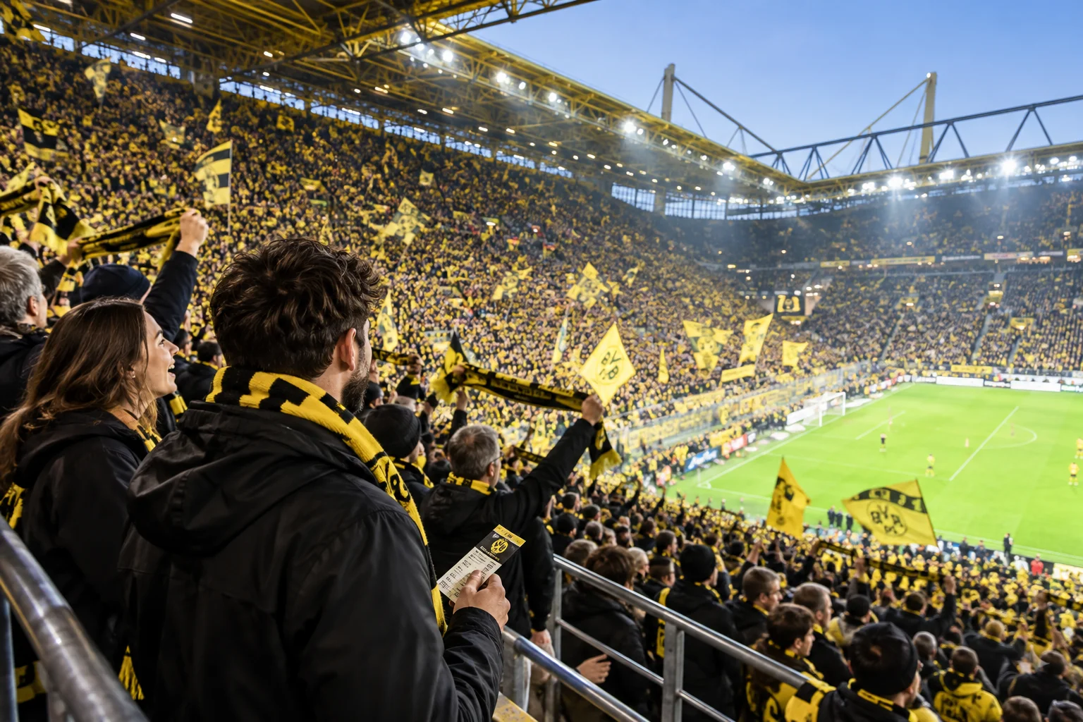 Borussia Dortmund supporters holding up yellow team scarves and tickets while standing in a packed Yellow Wall stand inside Signal Iduna Park during a match.