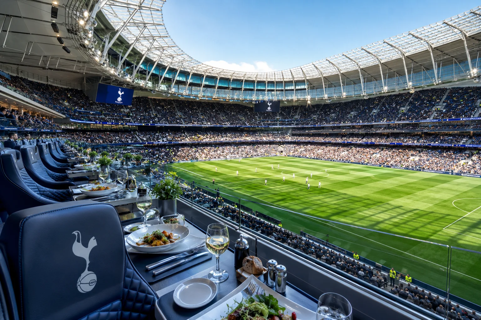 A formal VIP dining table set with white linen, champagne, and appetizers in an executive suite overlooking the empty pitch and red seats of Manchester United's Old Trafford stadium.