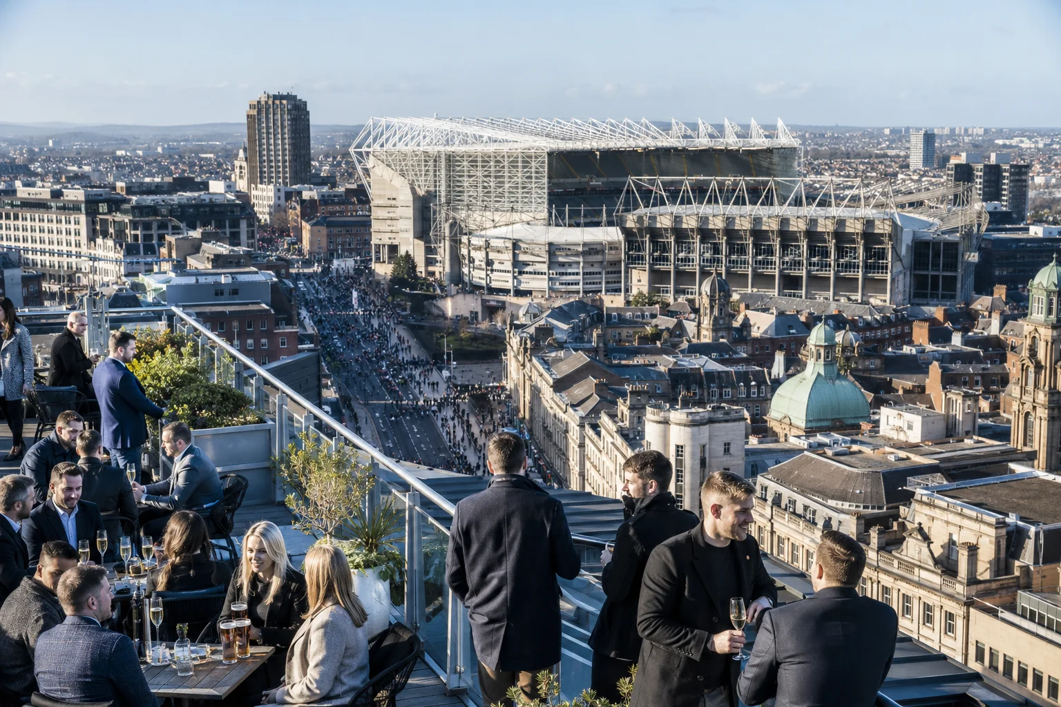 Guests interacting with staff in a modern, two-story luxury hospitality suite featuring gourmet food platters, drinks, comfortable seating, and wide views of a stadium football pitch during a game.