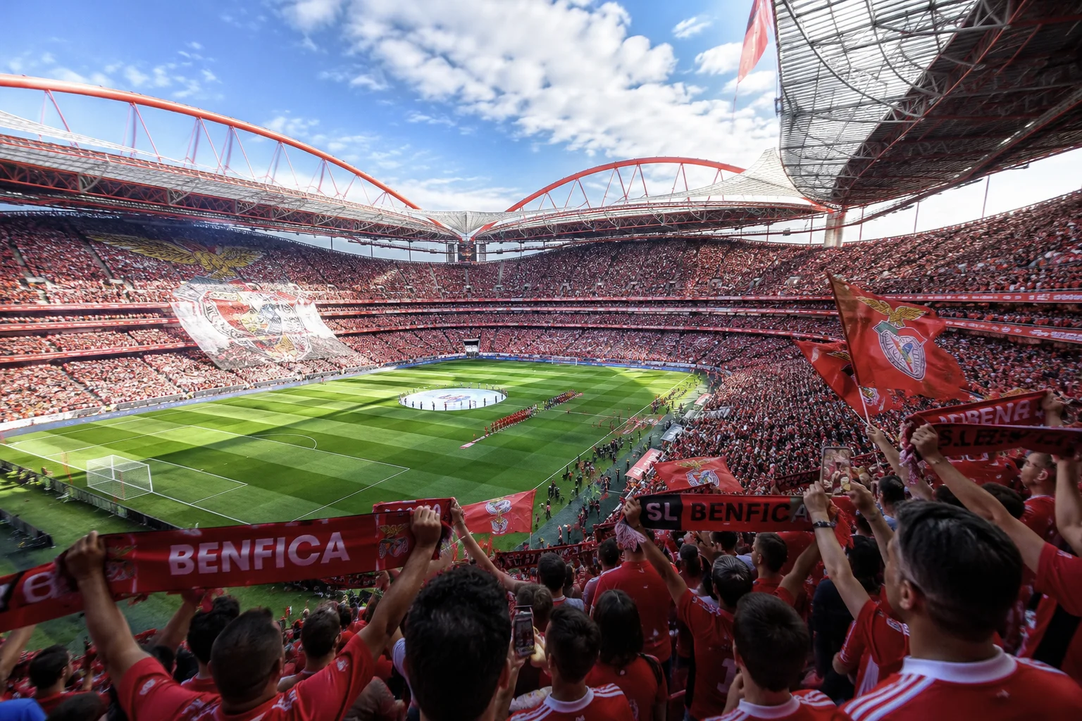 High-angle view of a full soccer stadium where fans wave red flags and stretch Benfica scarves across the stands as players line up on the pitch below.