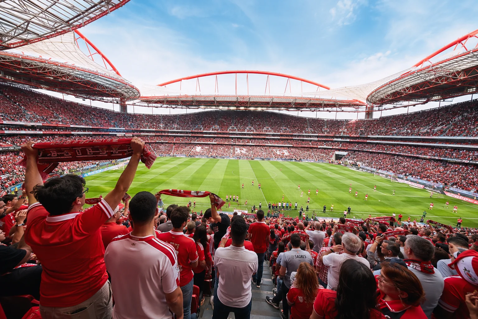 Wide interior view from the stands of a packed stadium during daytime as Benfica fans dressed in red hold up scarves overlooking the bright green pitch.
