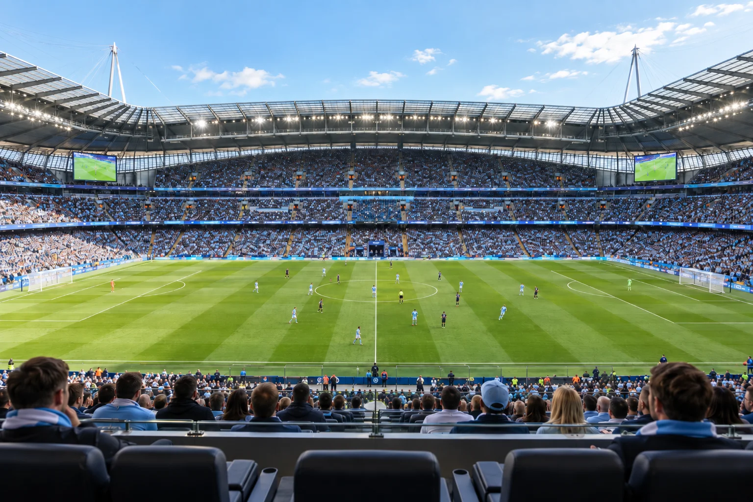 View of the football pitch centered from executive-style padded seats behind the lower stand barrier, showing players spread across the field.