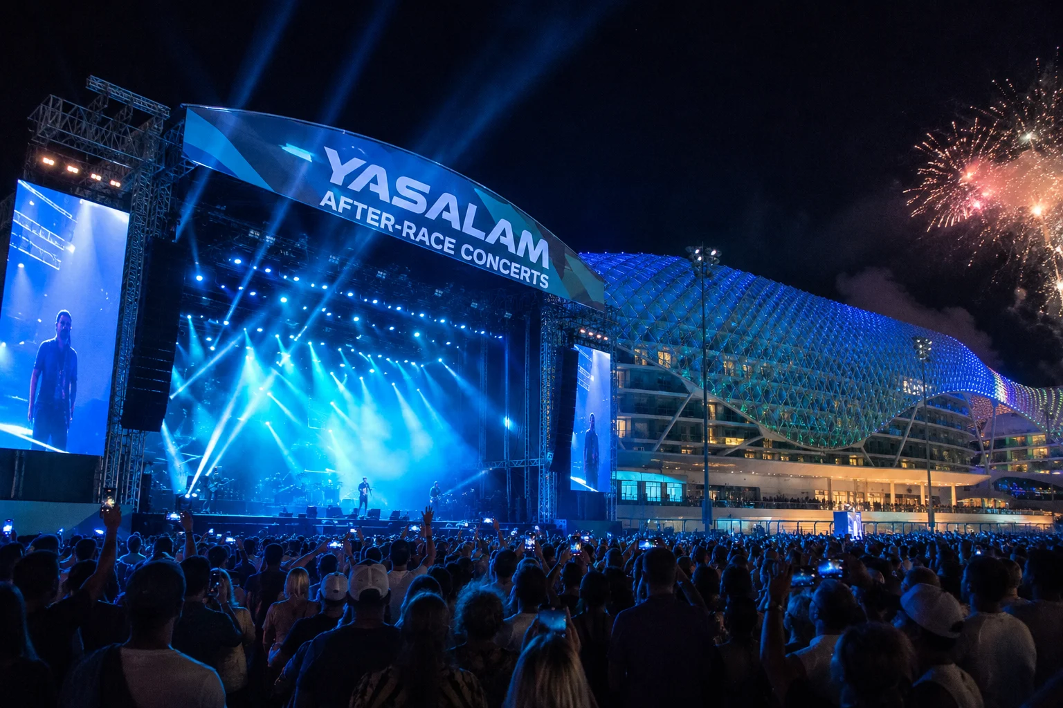 A outdoor night concert crowd gathered in front of a massive stage featuring blue lighting and screen displays beneath a banner reading "Yasalam After-Race Concerts," with fireworks exploding near the illuminated hotel structure in the background.