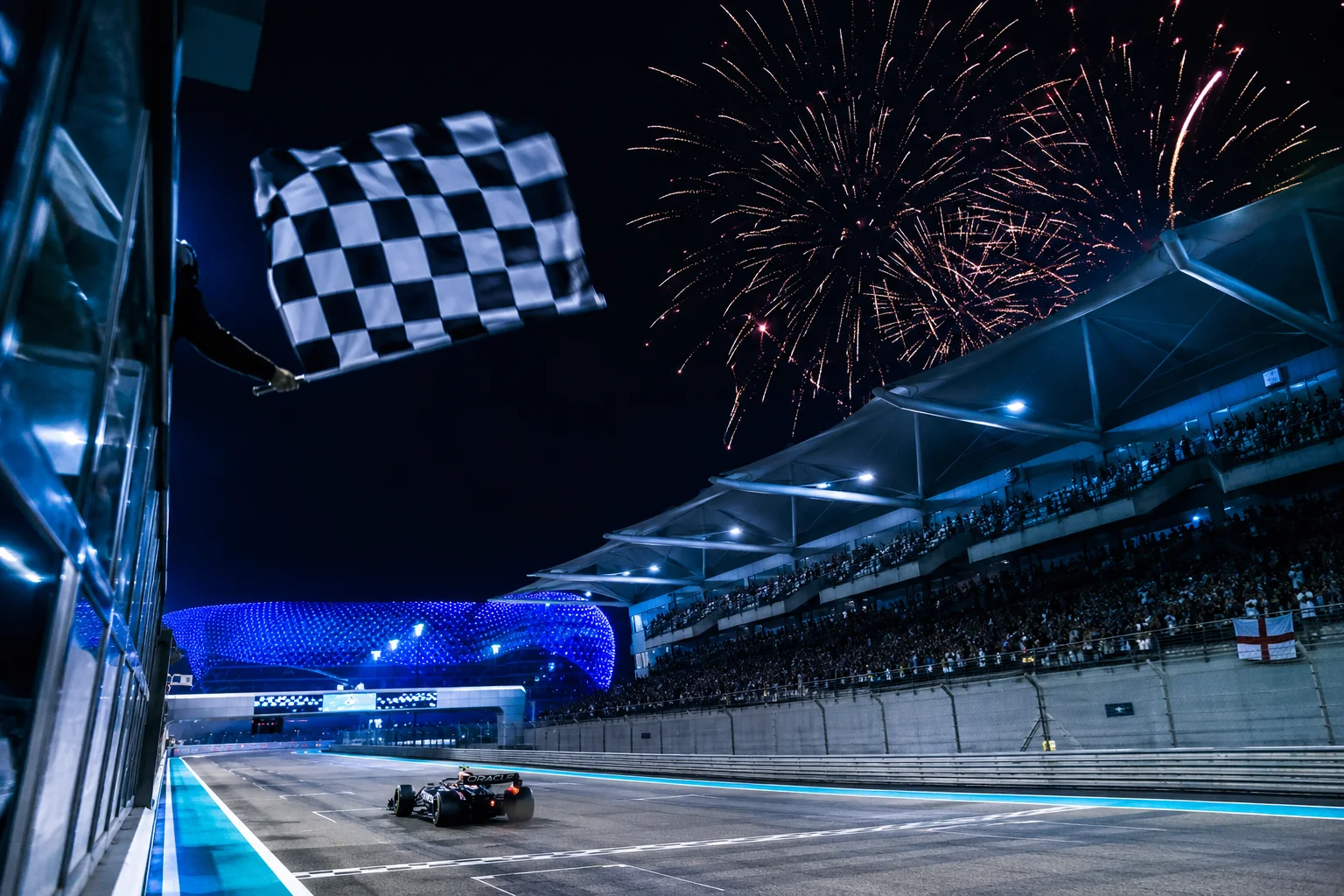 A dramatic night scene of a Formula 1 car crossing the finish line beneath a waved checkered flag, with spectator grandstands filled with fans on the right and vibrant fireworks bursting in the dark sky above.
