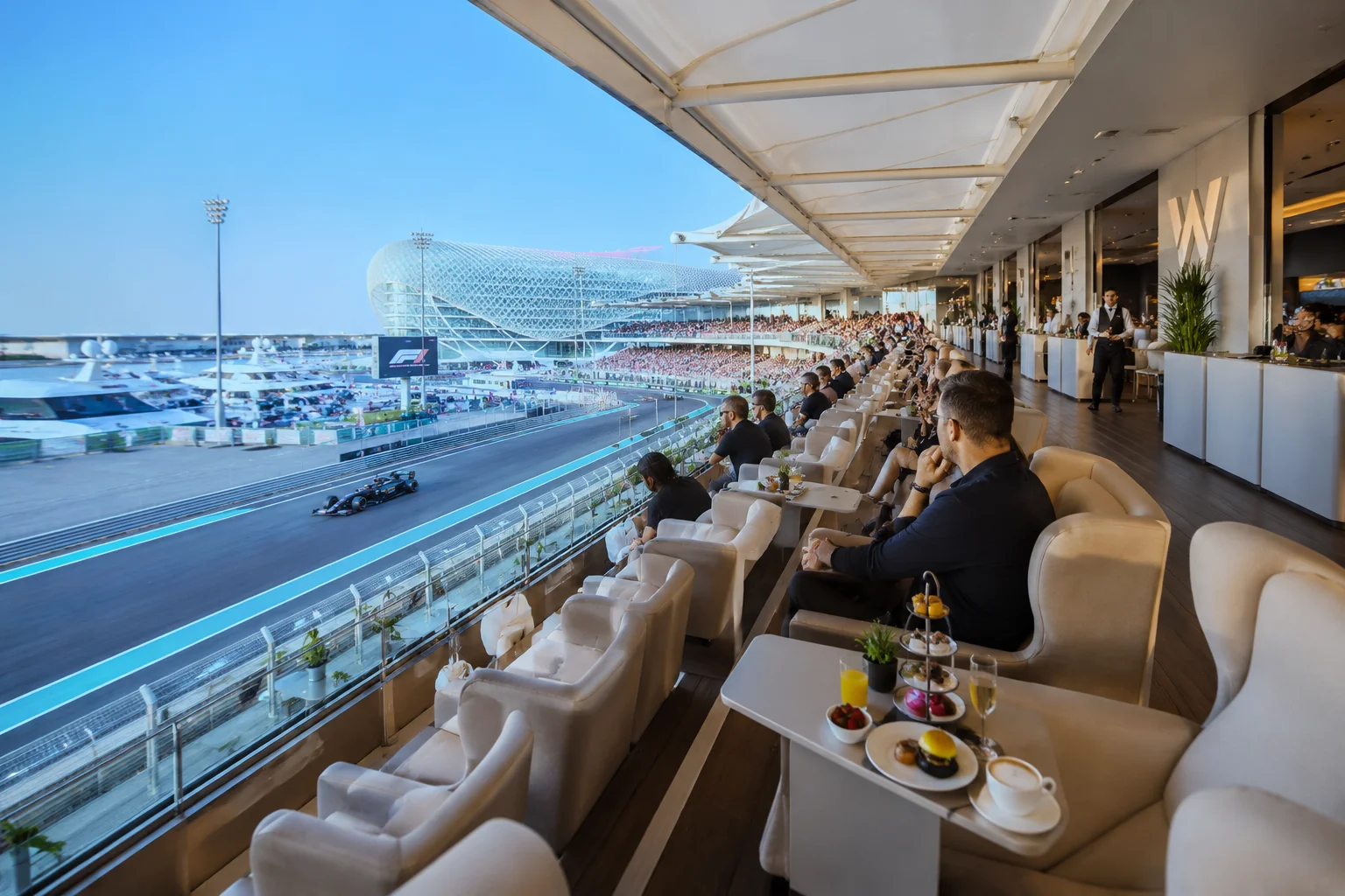 Guests seated in plush armchairs along a luxury hospitality suite balcony at the W Hotel, enjoying food and beverages at personal tables while taking in an elevated view of an F1 car racing along the track below.