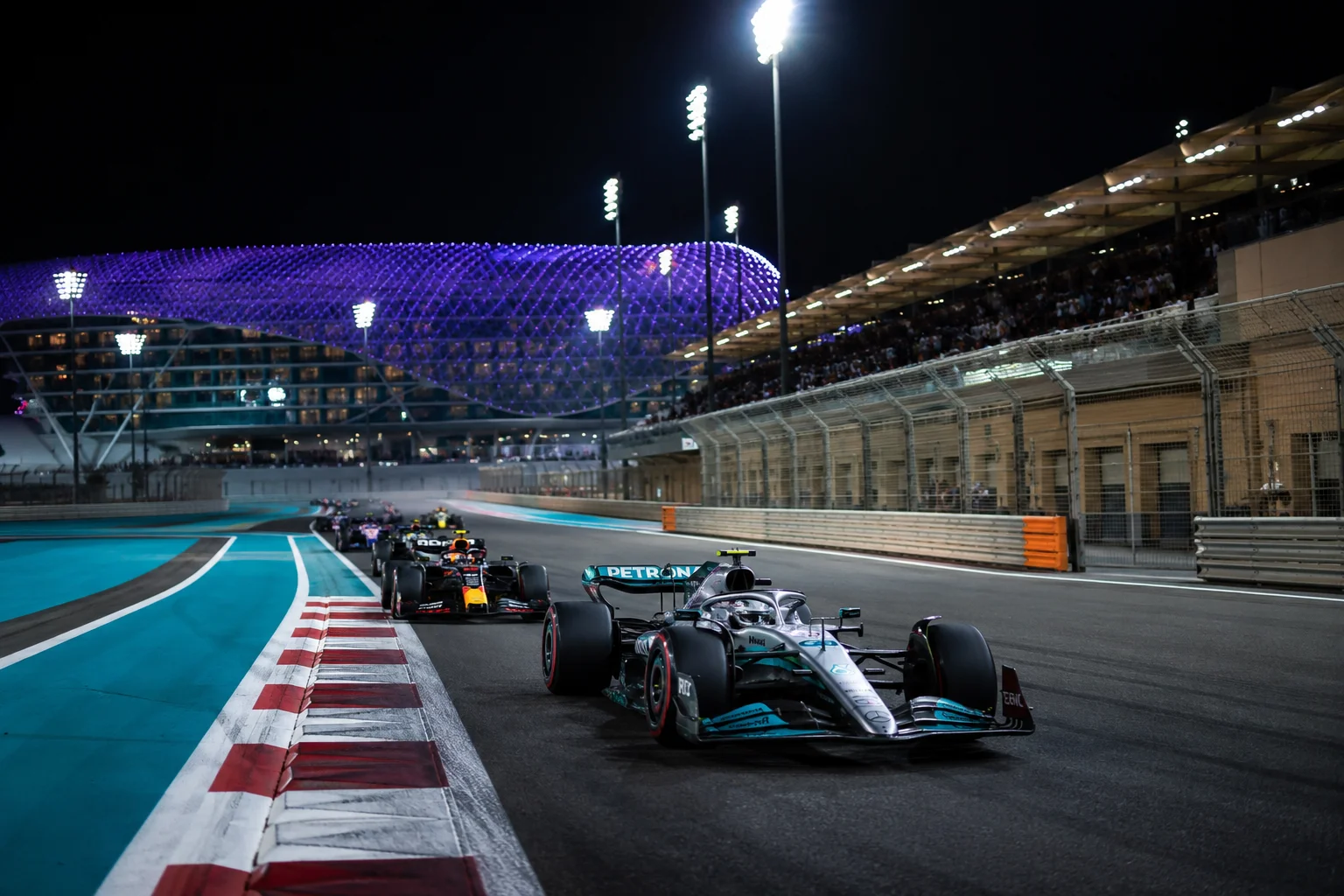 A night race action shot capturing a silver Mercedes Formula 1 car leading a line of competing race cars through a turn on the track, illuminated by bright stadium lights with the purple-lit grid structure of the hotel visible in the background.