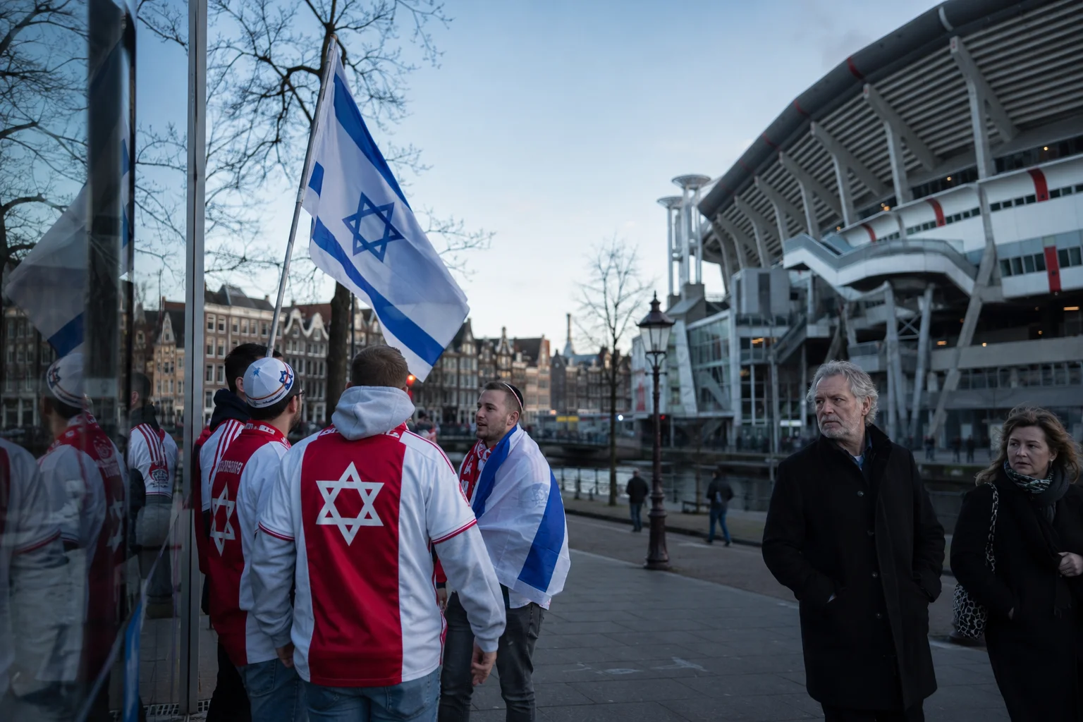 A group of young men wearing white and red football jerseys featuring the Star of David stand together on a city sidewalk near a canal, with one holding an Israeli flag. In the foreground to the right, a older man in a black coat and a woman walk past along the paved walkway, with a large modern stadium structure visible across the water in the background.