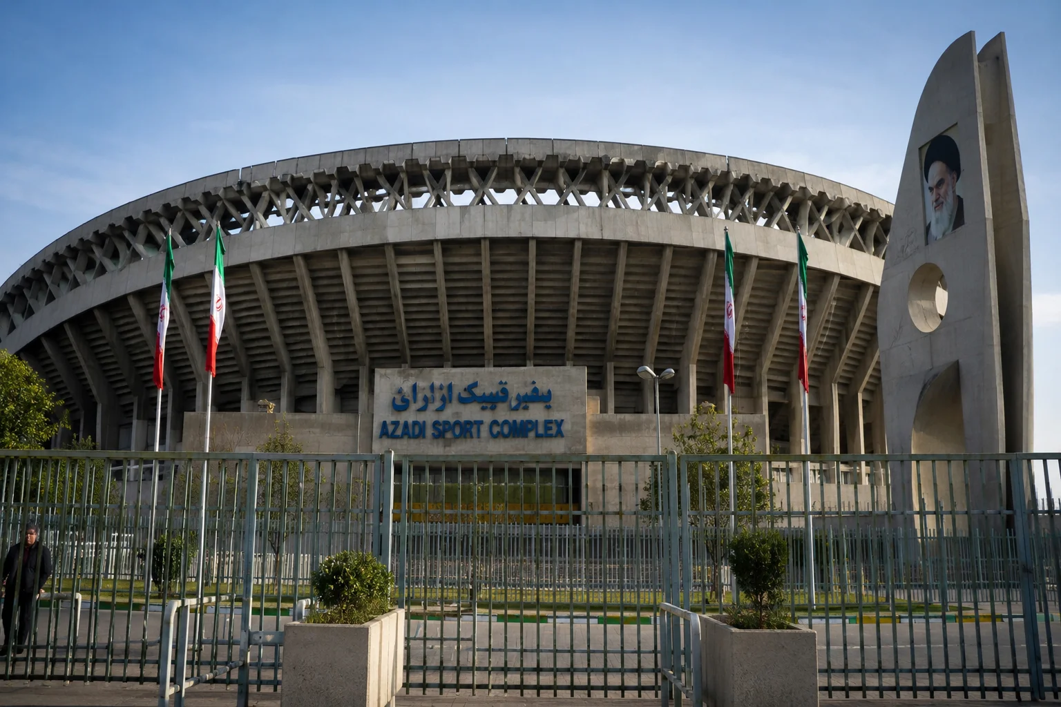 An exterior shot of the brutalist concrete Azadi Sport Complex stadium under a clear sky. A tall metal perimeter fence sits in the foreground, with several Iranian flags flying on poles behind it. Blue signage above the entrance displays text in both Persian script and English reading "AZADI SPORT COMPLEX," while a concrete pillar to the right features a portrait of Ayatollah Khomeini.