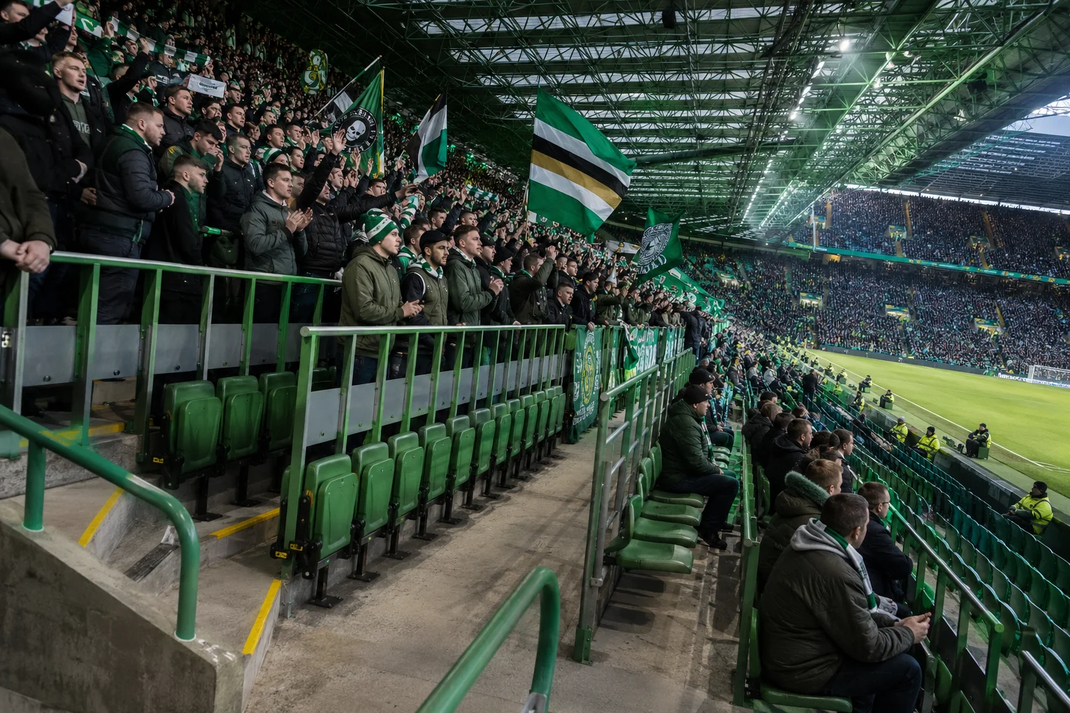 A view along the standing terrace section of a filled football stadium packed with active supporters. Fans dressed in green and black jackets clap, chant, and wave large green, white, and black striped flags. The stadium stands extend deep into the background under a green-tinted steel roof structure overlooking the bright green pitch.