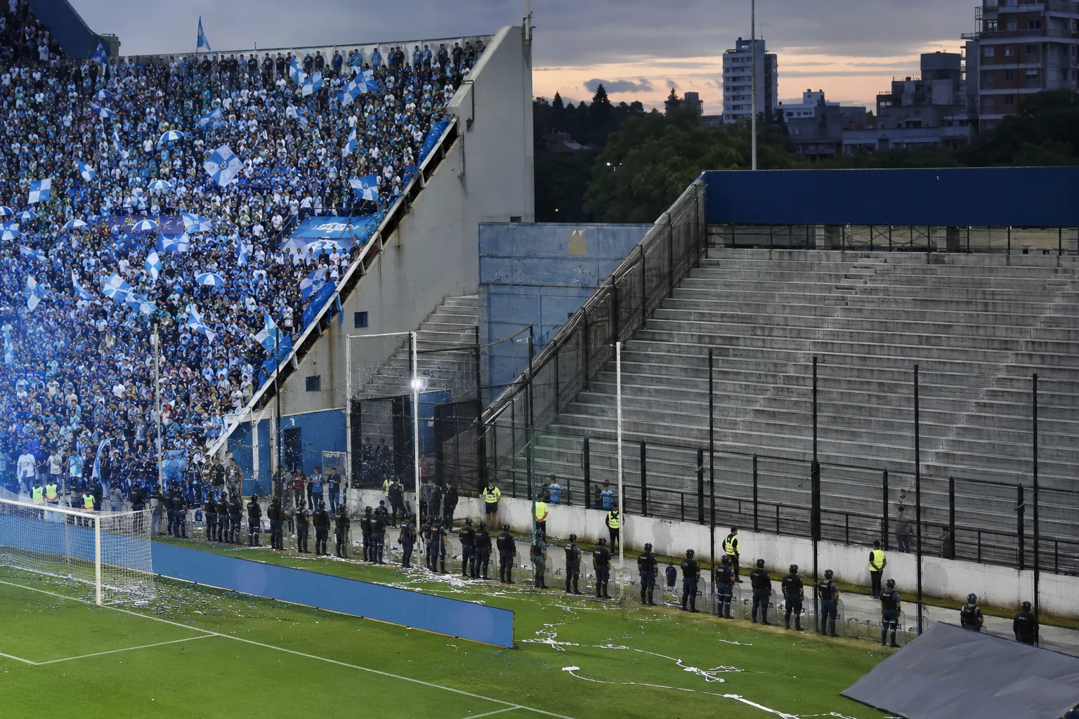 A wide shot inside a football stadium showing a steep section of grandstands on the left completely packed with cheering fans in light blue and white waving flags, while the adjacent right section of concrete stands remains entirely empty. A line of riot police in full gear stands along the perimeter track on the pitch below, creating a security barrier in front of the supporters.