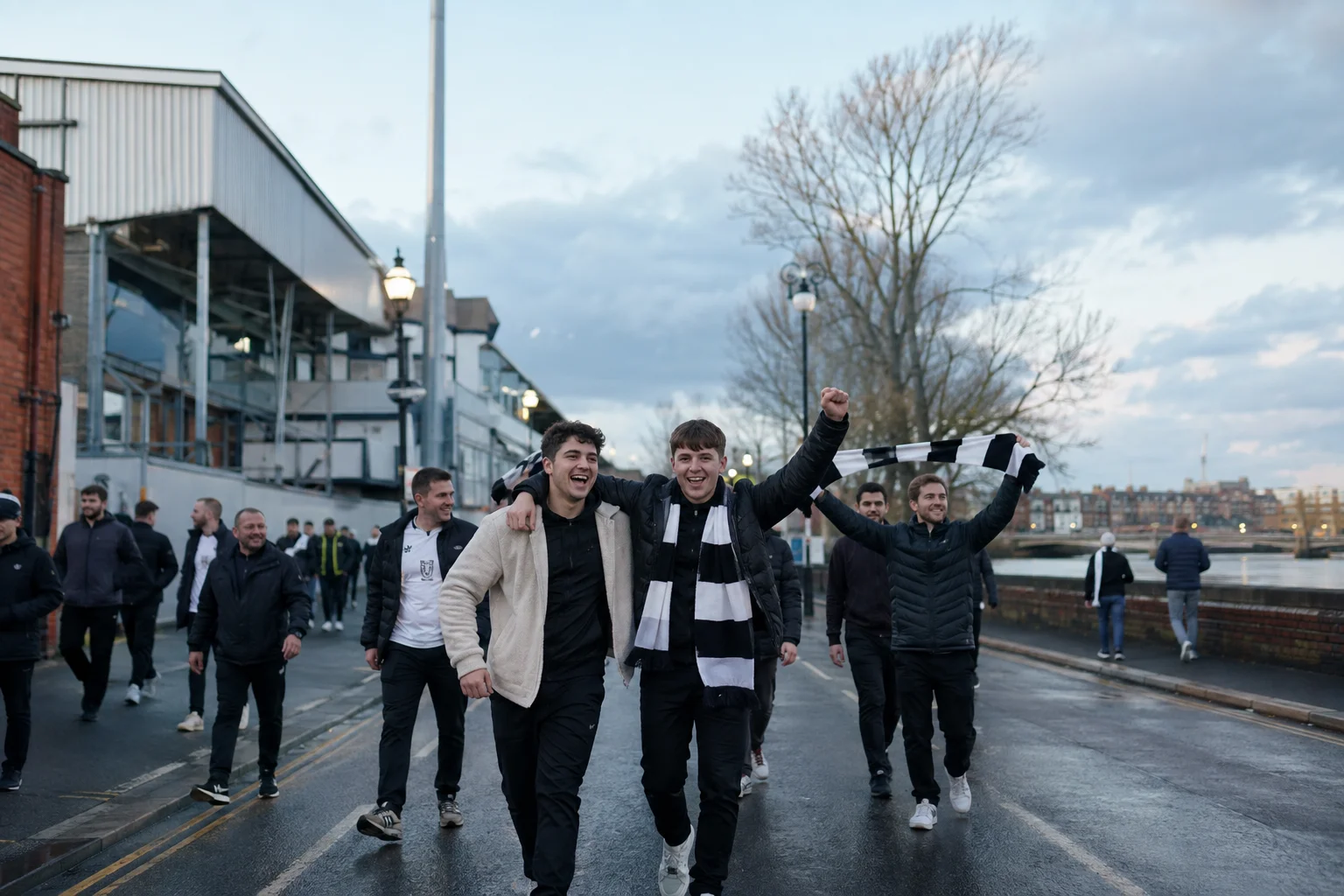 Happy young Fulham supporters cheering and holding up a team scarf while walking near Craven Cottage stadium.
