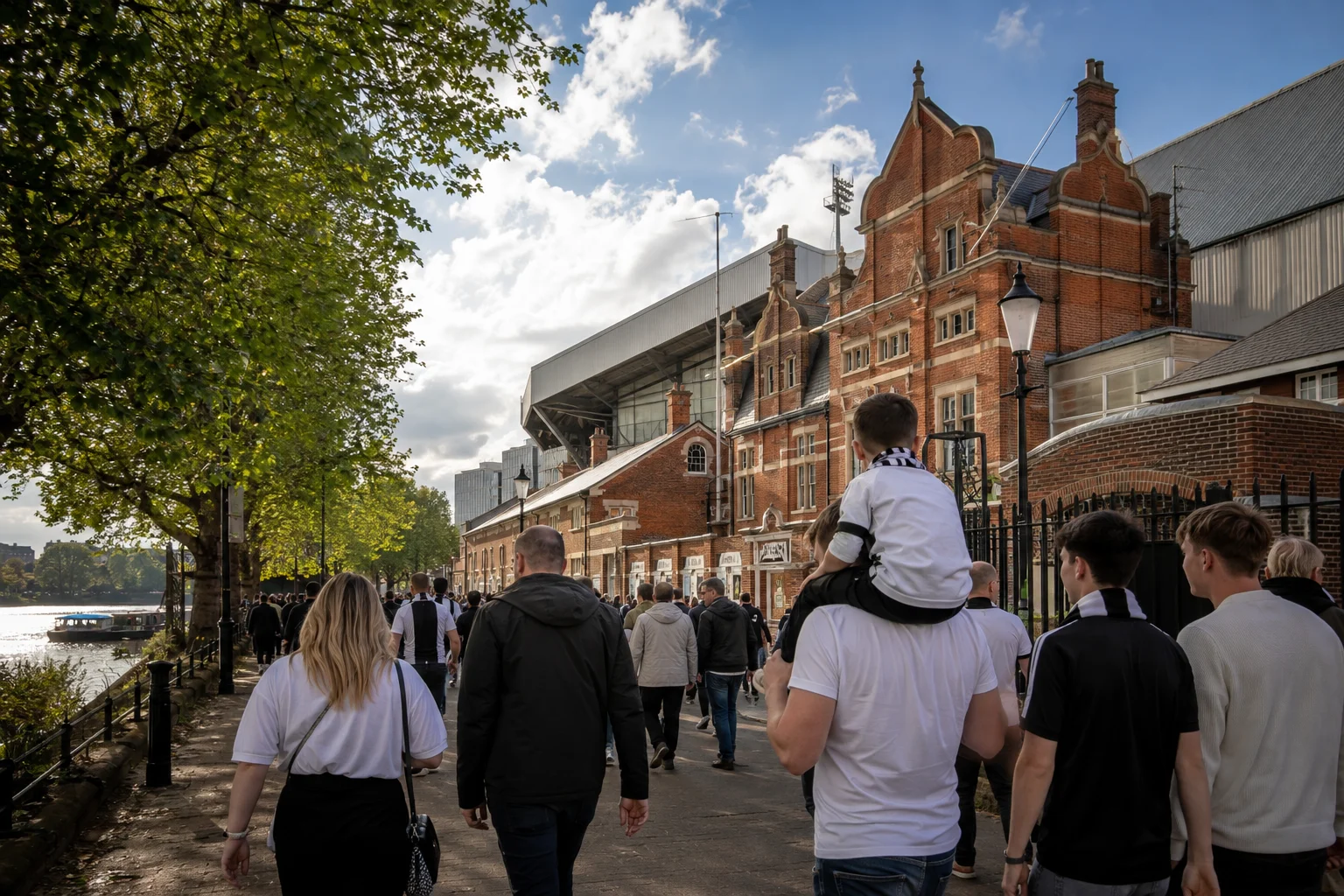 Fulham FC fans, including families and children, walking along the River Thames pathway outside Craven Cottage stadium on a matchday.