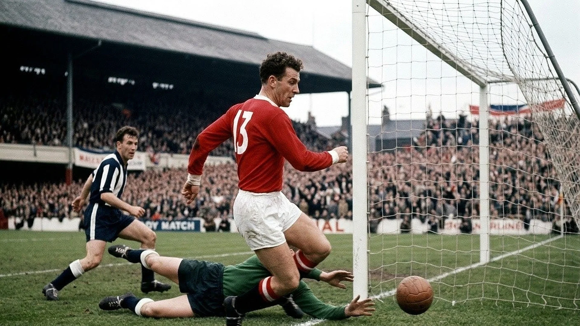 captures a dramatic moment right in front of the goal during a vintage football match. In the center, an attacker wearing a long-sleeved red jersey with the number 13 on the back and white shorts watches the play develop. Below him, a goalkeeper in a green jersey and dark shorts stretches out along the grass, extending his arms in an attempt to stop a classic brown leather football rolling across the goal line. Another outfield player in a vertically striped dark blue and white jersey runs toward the action from the left. In the background, a massive crowd of spectators packed tightly into covered grandstands watches the match closely.