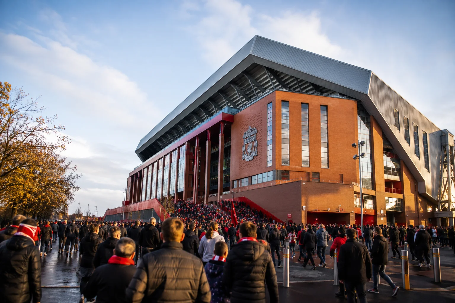 Crowd of football fans wearing red scarves walking toward the Main Stand exterior at Anfield Stadium on matchday.