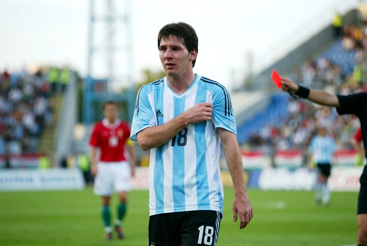 شاب ليونيل ميسي يبدو عاطفيًا في قميص المنتخب الأرجنتيني رقم 18 بينما يحمل الحكم بطاقة حمراء في المقدمة خلال debut الدولي.