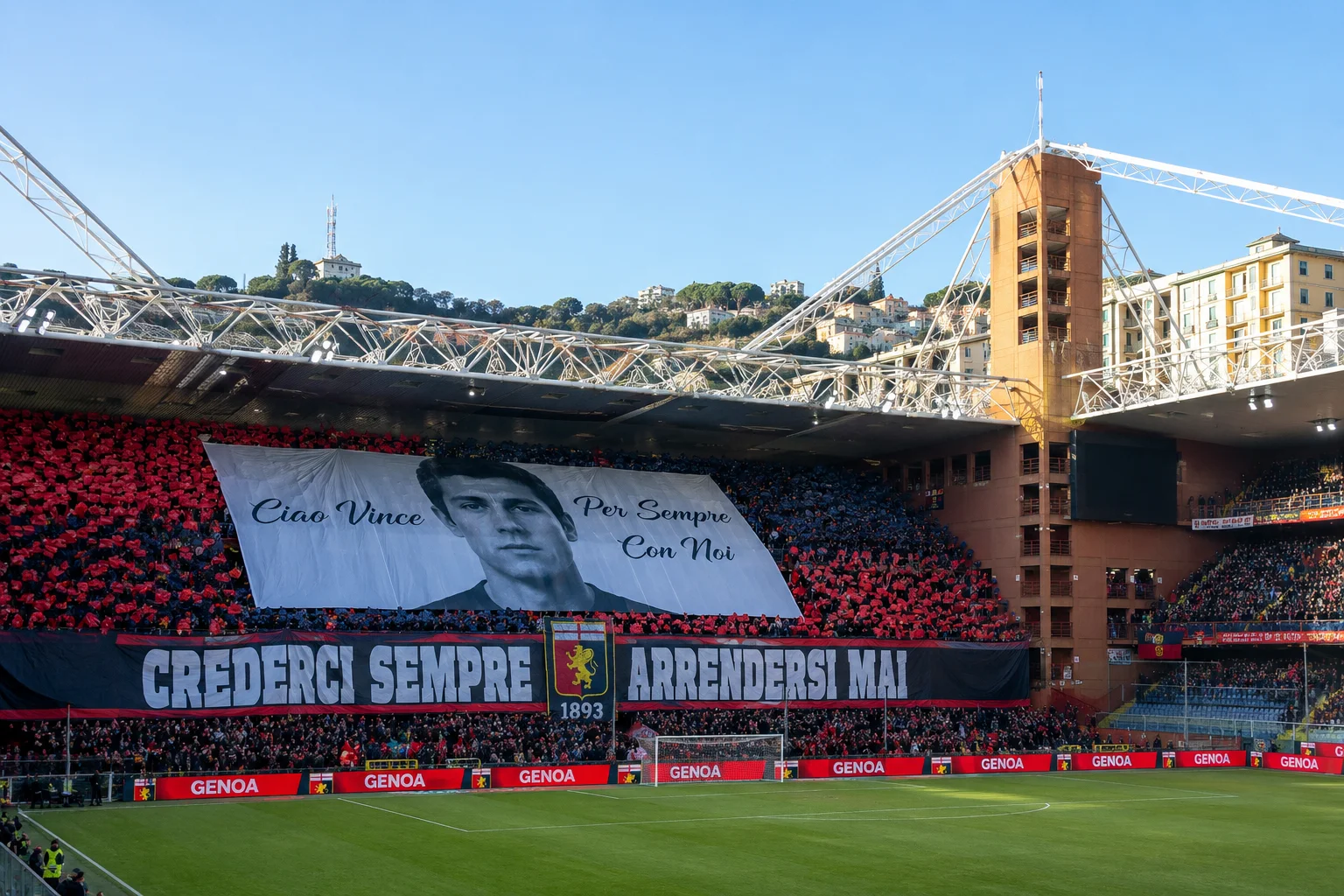 A wide-angle view of the interior of Genoa's Luigi Ferraris Stadium filled with fans. A massive white memorial banner hangs across the main stand displaying a portrait of a young man along with the Italian text "Ciao Vince Per Sempre Con Noi". Below it, a large black banner reads "CREDERCI SEMPRE ARRENDERSI MAI" with Genoa's coat of arms in the center, set above the bright green pitch.