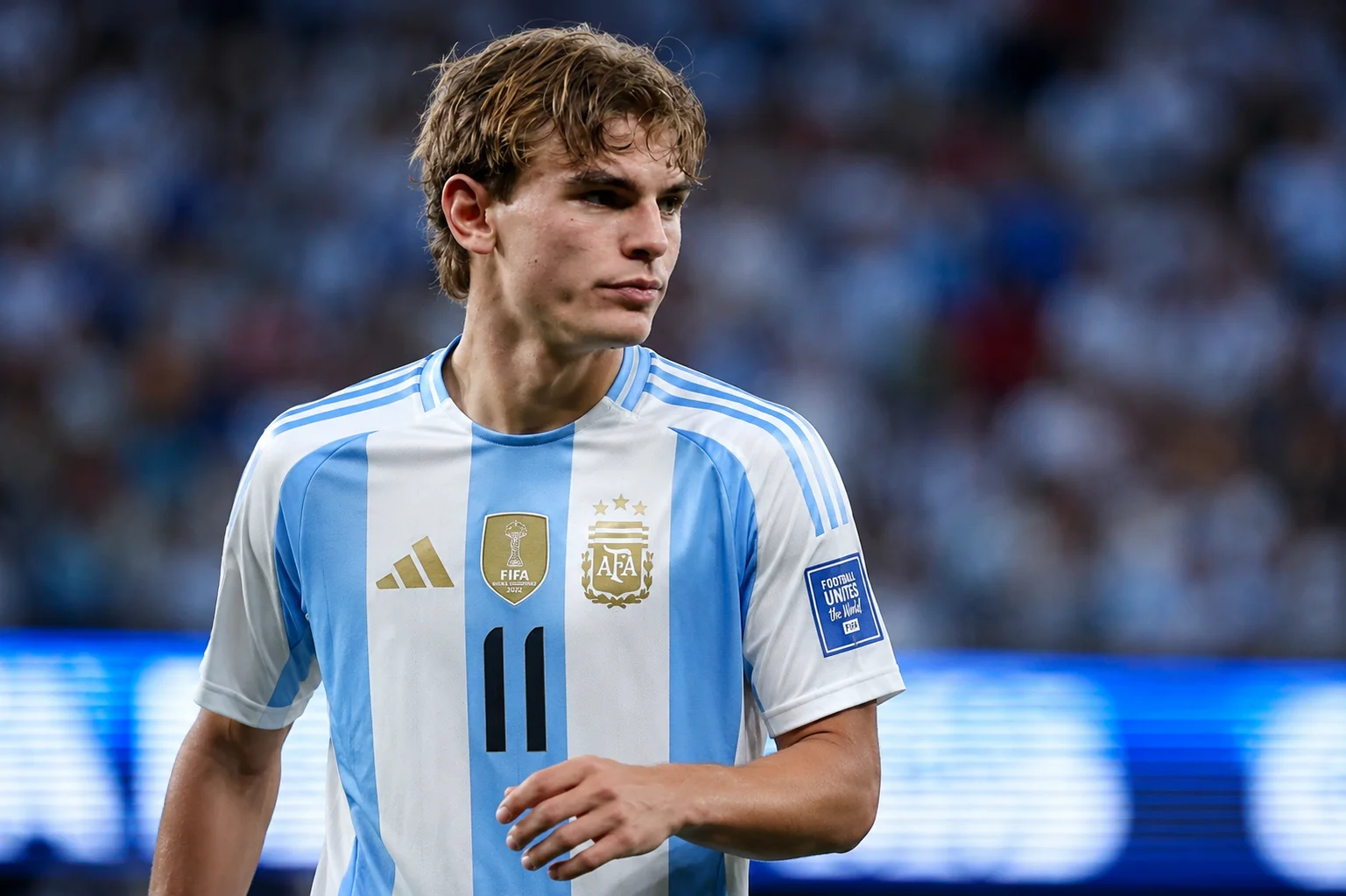 لقطة حركية للاعب الوسط الأرجنتيني نيكو باز يتتبع اللعب على الملعب. يتم تصويره من الصدر إلى الأعلى، في وضع حركة مع رأسه مائل نحو اللعب. يرتدي القميص الكلاسيكي المائل الأزرق والأبيض والذي يتضمن الشعار الذهبي للاتحاد الأرجنتيني لكرة القدم. تُظهر الخلفية العشب الأخضر الكثيف وأبراج الهيكل المموهة لملعب كبير في الخلفية.