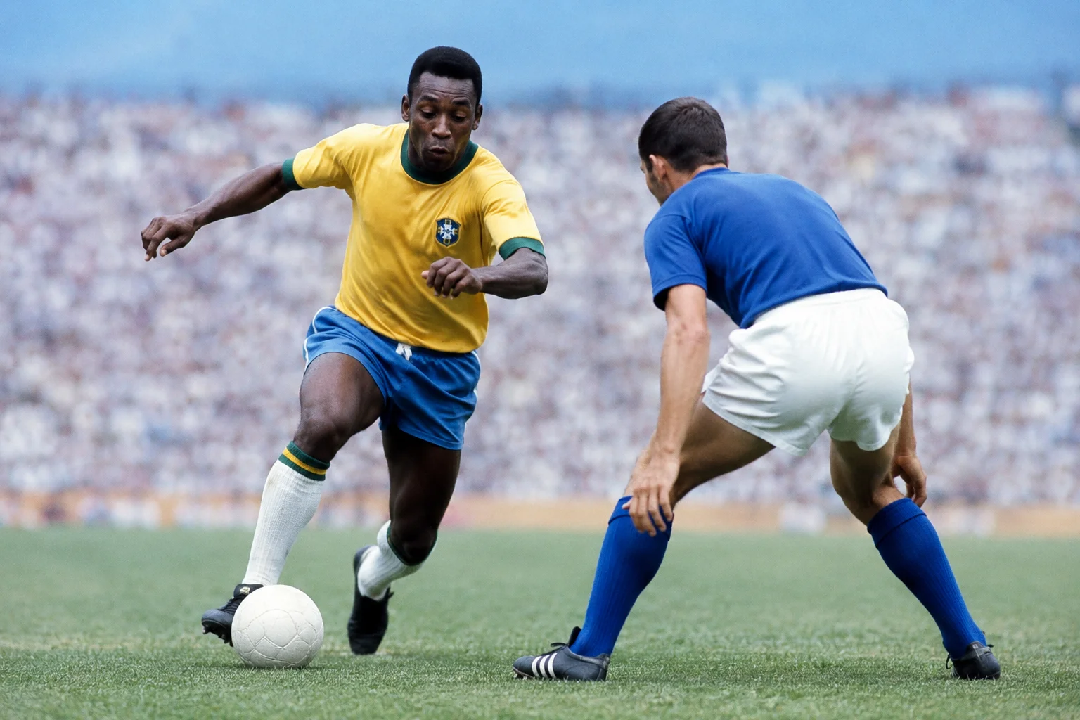 A vintage action shot shows the legendary Brazilian soccer player Pelé wearing his iconic yellow and green national team jersey and blue shorts as he drives forward with a white soccer ball on a lush green pitch. He is intensely focused, utilizing a dynamic body feint to outmaneuver an opposing defender dressed in a blue jersey and white shorts who is crouched and anticipating his next move. A massive, packed stadium crowd creates a blurred backdrop to the encounter.