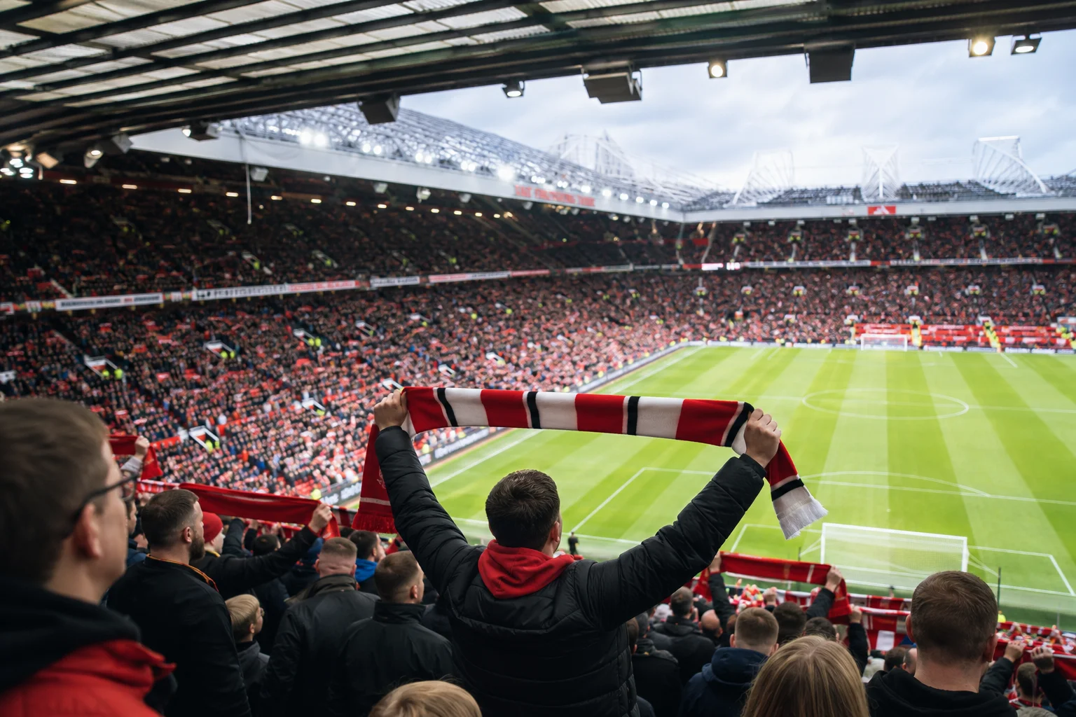 A view from the packed stands of a massive stadium where a fan in the foreground holds a stretched red, white, and black scarf above their head, looking out over the bright green soccer pitch.