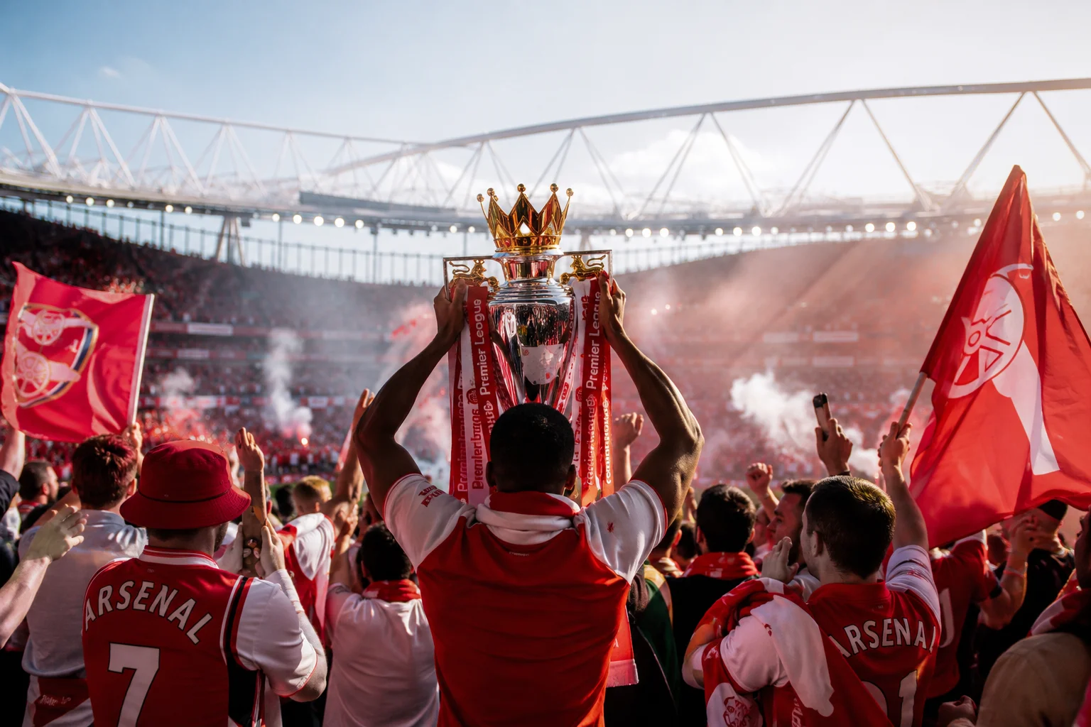 Seen from behind, a soccer player in a red and white Arsenal jersey lifts the silver, gold-crowned Premier League trophy high into the air, surrounded by a roaring crowd, red smoke flares, and waving team flags.