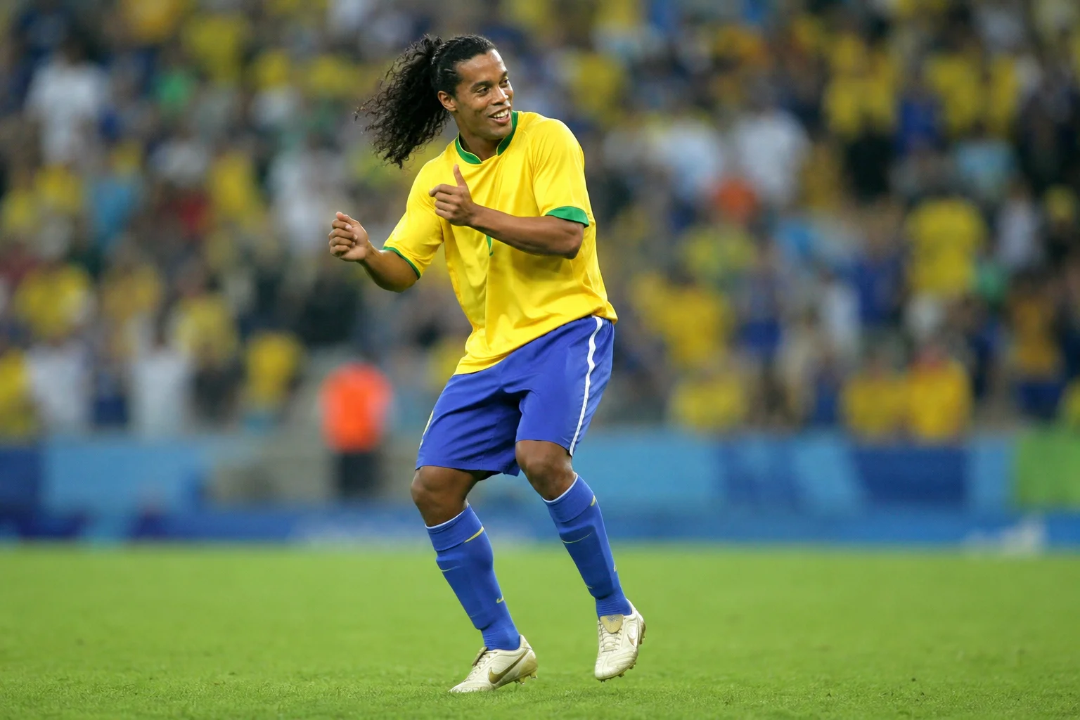 Brazilian soccer star Ronaldinho, wearing the classic yellow national team jersey and blue shorts, smiles brightly on a green field as he performs a joyful, rhythmic dance celebration. His long, curly hair bounces mid-motion, and he holds his hands up with thumbs raised, embodying the playful "Joga Bonito" spirit. The background features a vibrant, softly blurred stadium filled with spectators wearing yellow.
