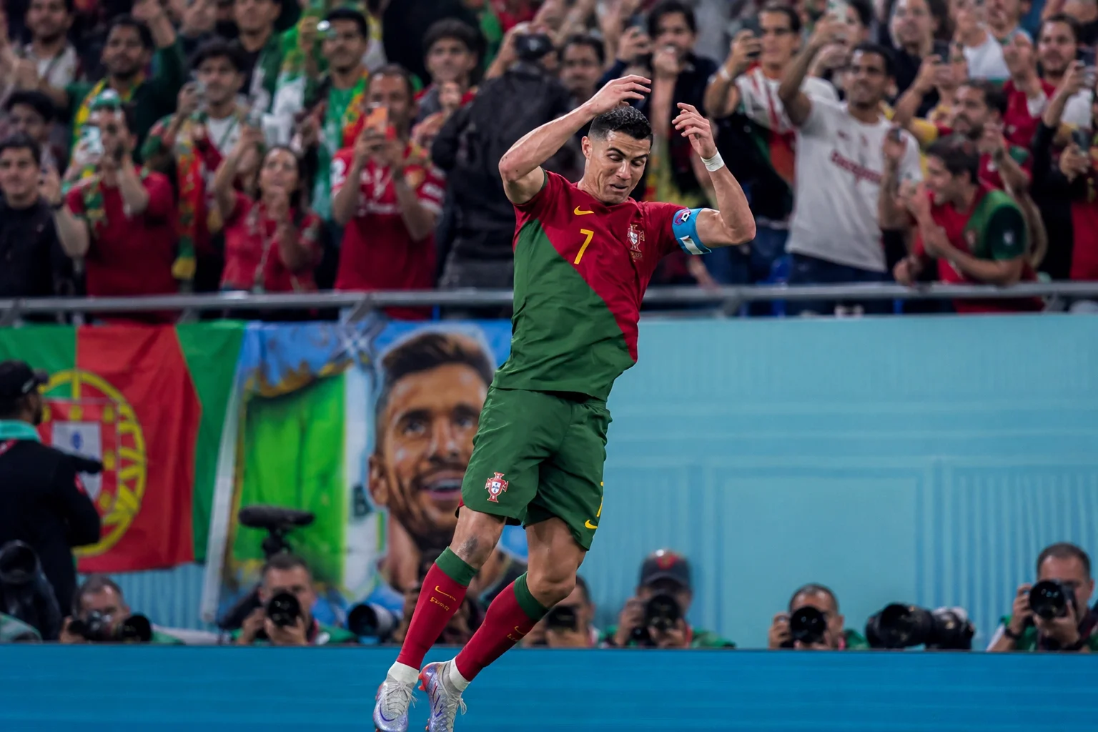 لاعب كرة قدم محترف كريستيانو رونالدو في الهواء على الملعب، يؤدي احتفاله المميز "سيوو". يرتدي قميص البرتغال الأحمر والأخضر مع الرقم 7، وشورتات مطابقة، وعصابة كابتن زرقاء، مع ذراعيه مرفوعتين ومرفقيه منحنين. في الخلفية الضبابية، تشجع حشود متجمعة في الاستاد، ويحمل المشجعون هواتفهم الذكية، وصف المصورون الذين يحملون عدسات كبيرة على حافة الملعب.