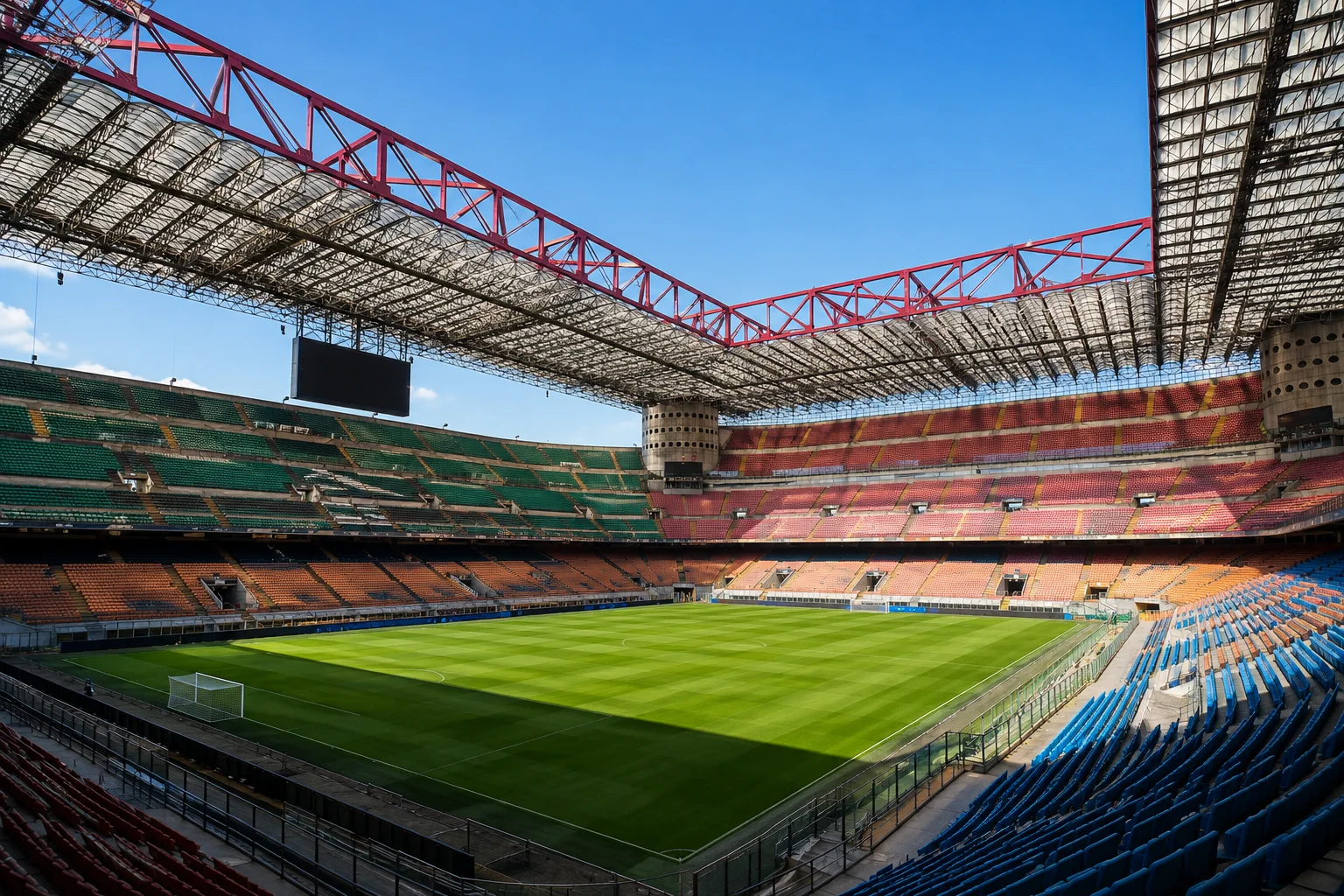 A wide-angle, pitch-level view of an empty San Siro stadium on a bright, clear day. The vibrant green grass field is framed by tiers of red, orange, green, and blue spectator seating surrounding the pitch. Dominating the upper structure is the iconic red steel roof truss framework attached to the stadium's distinctive cylindrical concrete towers.