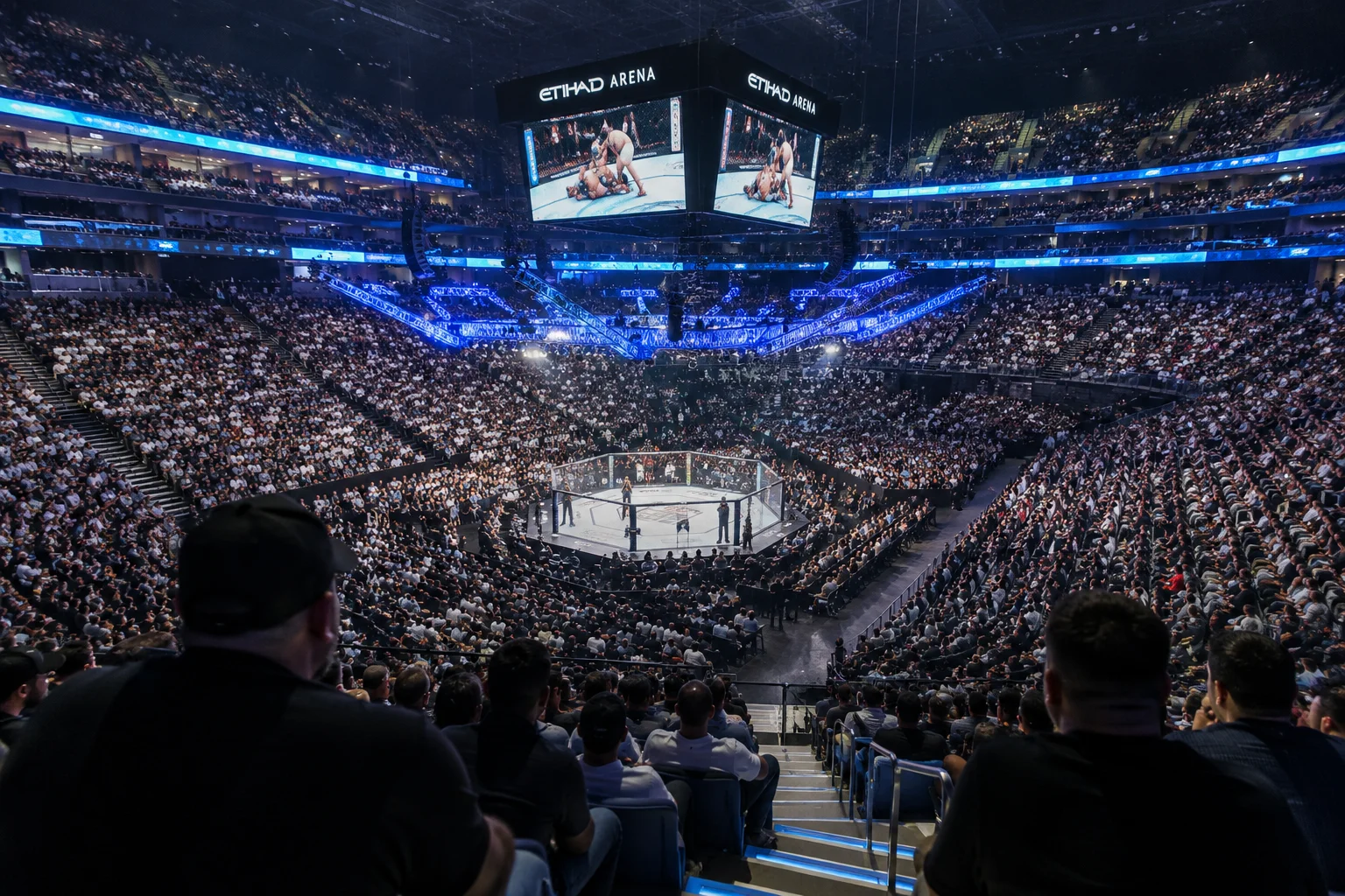 زاوية داخلية واسعة من الطبقة العليا لأرينا الاتحاد خلال حدث UFC مباشر، تظهر استاد مليء بالمشجعين يحيطون بمربع الضوء مع شاشات فيديو متعددة فوق الرأس تعرض المقاتلين أثناء العمل.
